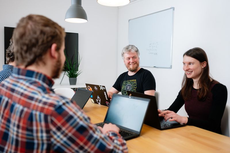 Drei Personen arbeiten gemeinsam mit Laptops in einem Besprechungsraum und tauschen sich über ein Projekt aus.